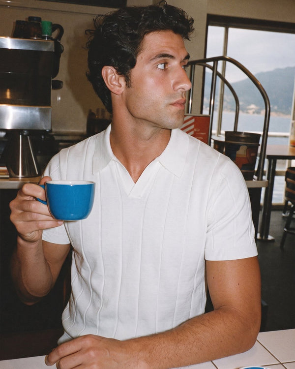 A man in a white knit polo having coffee by the water